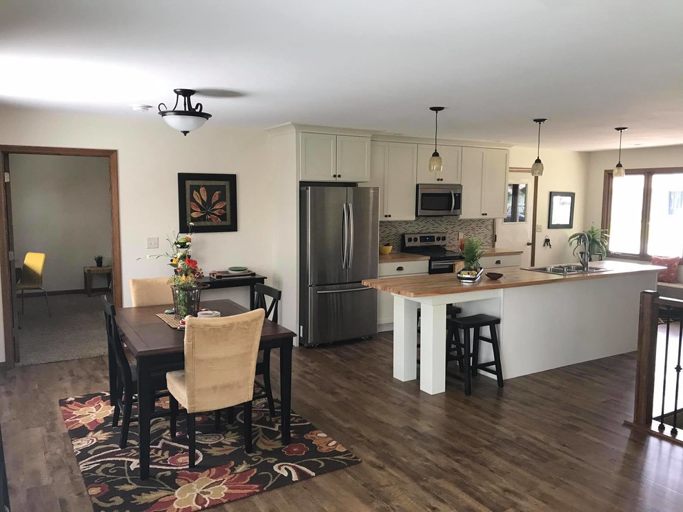 Example of a mid-sized transitional single-wall vinyl floor and brown floor open concept kitchen design with recessed-panel cabinets, white cabinets, wood countertops, glass tile backsplash, an island, a drop-in sink, gray backsplash, stainless steel appliances and brown countertops