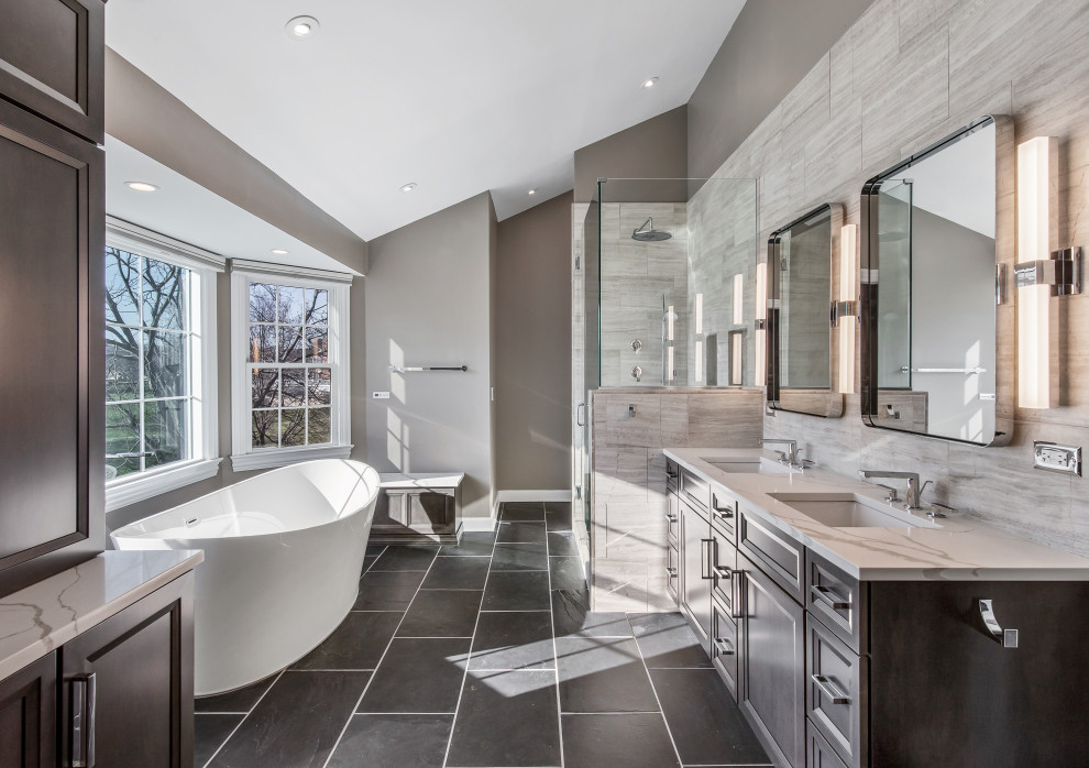 This is an example of a large contemporary master bathroom in Chicago with flat-panel cabinets, medium wood cabinets, a freestanding tub, an open shower, a one-piece toilet, gray tile, limestone, grey walls, slate floors, an undermount sink, engineered quartz benchtops, black floor, a hinged shower door, white benchtops, a niche, a double vanity, a built-in vanity and vaulted.