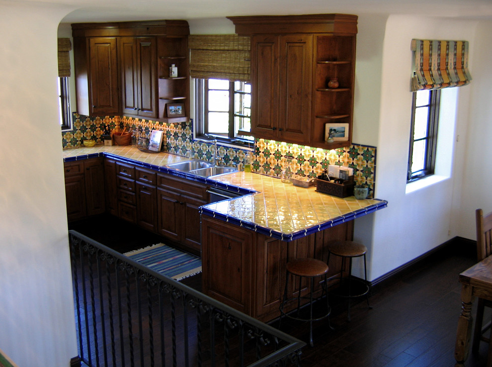 Inspiration for a small mediterranean u-shaped eat-in kitchen in Santa Barbara with a double-bowl sink, raised-panel cabinets, medium wood cabinets, tile benchtops, ceramic splashback, stainless steel appliances, dark hardwood floors, a peninsula, multi-coloured splashback, brown floor and yellow benchtop.