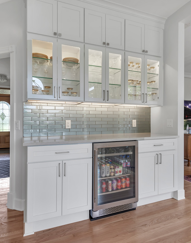This is an example of a transitional single-wall home bar in Detroit with no sink, shaker cabinets, white cabinets, quartz benchtops, grey splashback, subway tile splashback, medium hardwood floors, brown floor and grey benchtop.