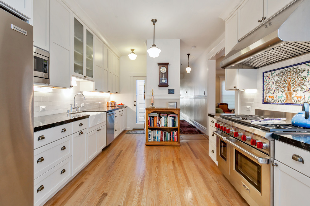 This is an example of a large country galley separate kitchen in San Francisco with a farmhouse sink, shaker cabinets, white cabinets, granite benchtops, white splashback, subway tile splashback, stainless steel appliances, light hardwood floors, with island and beige floor.