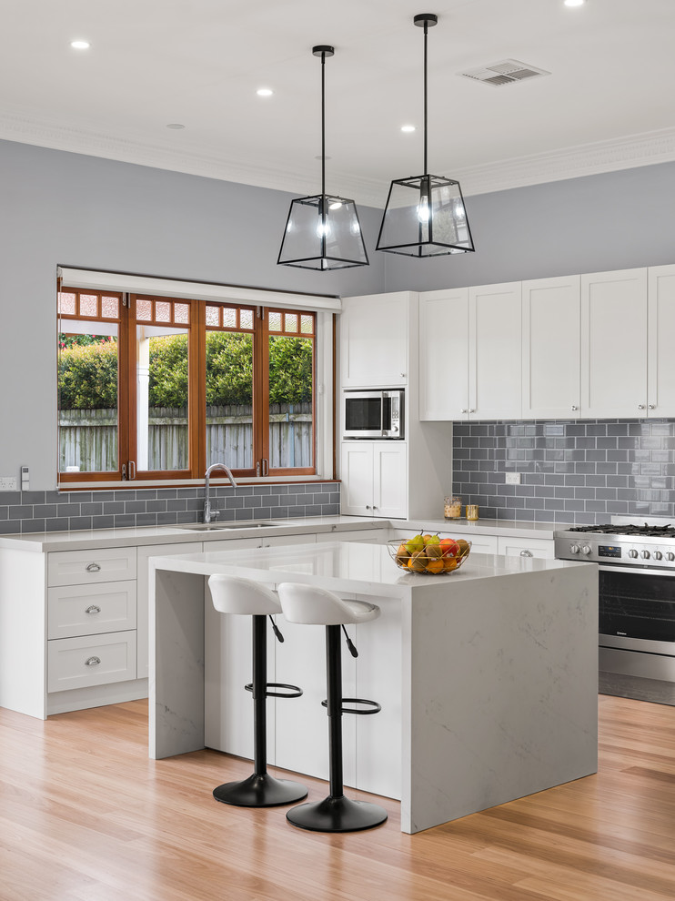Inspiration for a mid-sized traditional u-shaped open plan kitchen in Sydney with an undermount sink, shaker cabinets, white cabinets, quartz benchtops, grey splashback, ceramic splashback, stainless steel appliances, light hardwood floors, with island, brown floor and grey benchtop.
