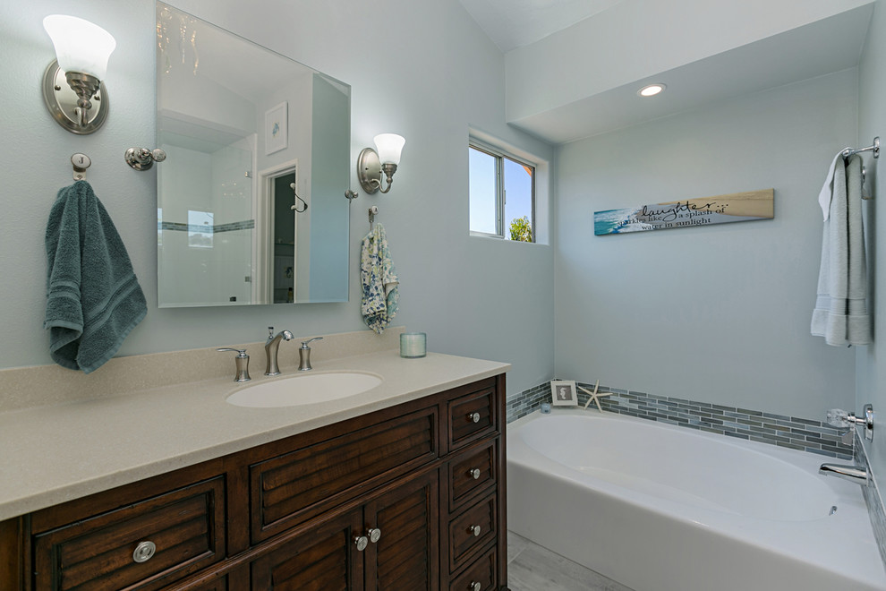 This is an example of a large beach style master bathroom in San Diego with furniture-like cabinets, medium wood cabinets, a corner tub, a corner shower, a two-piece toilet, gray tile, porcelain tile, grey walls, porcelain floors, a drop-in sink, granite benchtops, grey floor and a hinged shower door.
