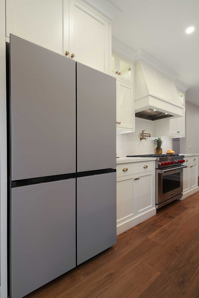 Large contemporary l-shaped eat-in kitchen in San Francisco with a farmhouse sink, shaker cabinets, white cabinets, solid surface benchtops, white splashback, subway tile splashback, stainless steel appliances, medium hardwood floors, with island, brown floor and white benchtop.