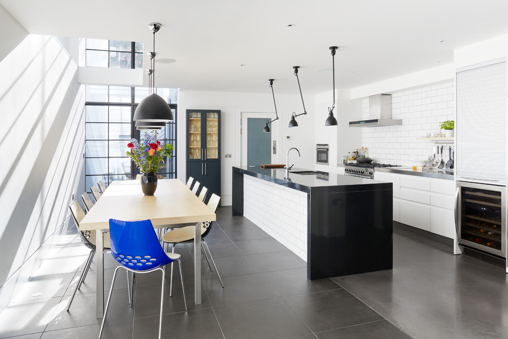 This is an example of a large contemporary single-wall eat-in kitchen in London with flat-panel cabinets, stainless steel benchtops, white splashback, subway tile splashback, stainless steel appliances, concrete floors and with island.