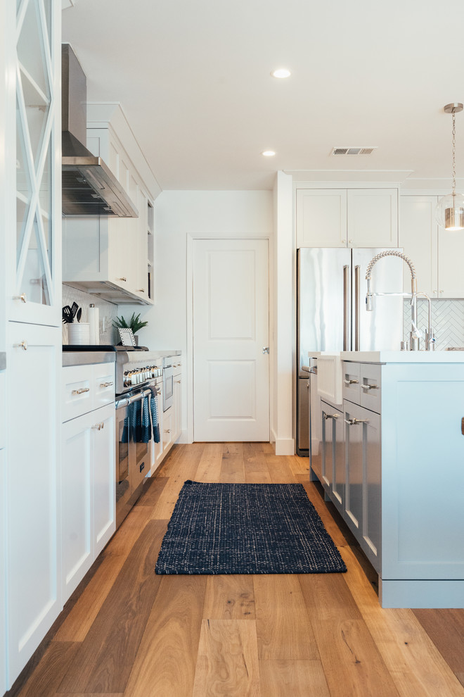 Photo of a transitional l-shaped kitchen in Los Angeles with a farmhouse sink, recessed-panel cabinets, white cabinets, stainless steel appliances, medium hardwood floors, with island, brown floor and white benchtop.