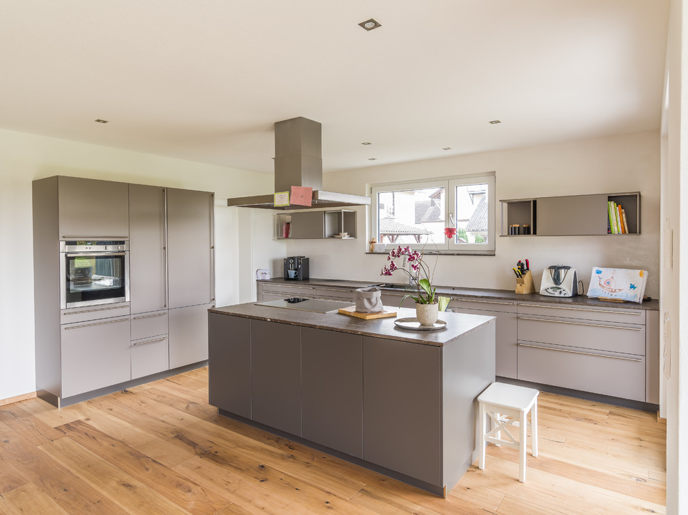 Photo of a large contemporary galley eat-in kitchen in Other with flat-panel cabinets, grey cabinets, white splashback, stainless steel appliances, medium hardwood floors, with island, an undermount sink, granite benchtops, limestone splashback, brown floor and brown benchtop.