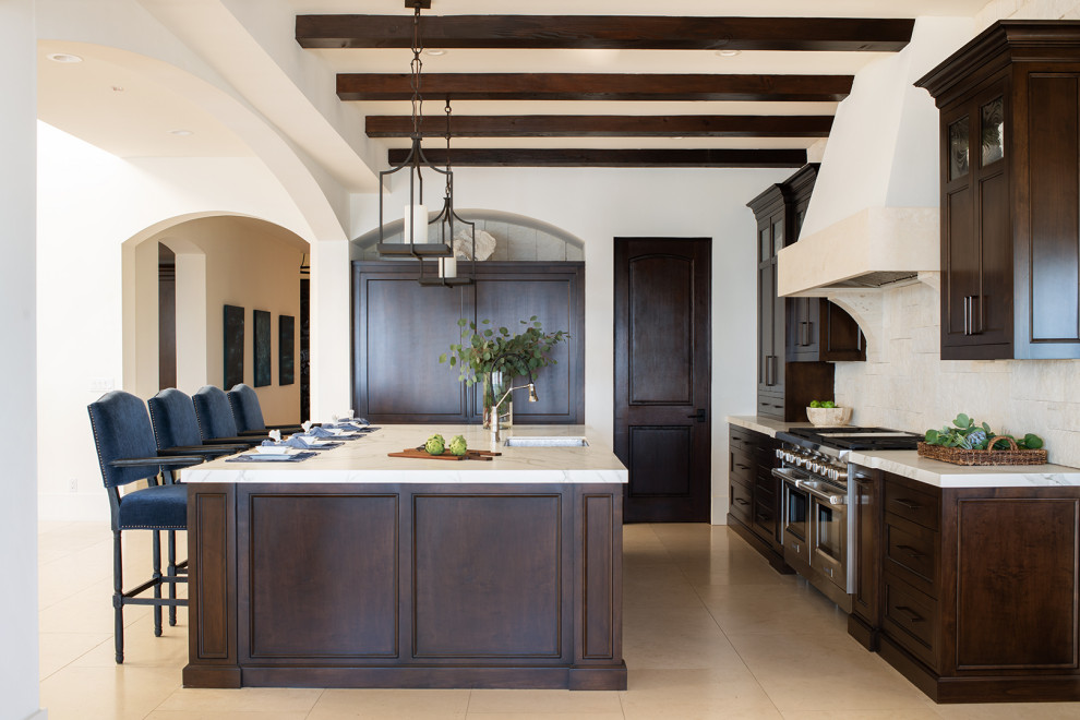 Photo of a large mediterranean galley open plan kitchen in Los Angeles with an undermount sink, recessed-panel cabinets, dark wood cabinets, solid surface benchtops, beige splashback, limestone splashback, panelled appliances, limestone floors, with island, beige floor, beige benchtop and exposed beam.