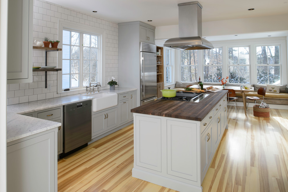 Inspiration for a mid-sized transitional l-shaped open plan kitchen in New York with a farmhouse sink, shaker cabinets, grey cabinets, wood benchtops, white splashback, subway tile splashback, stainless steel appliances, light hardwood floors and with island.