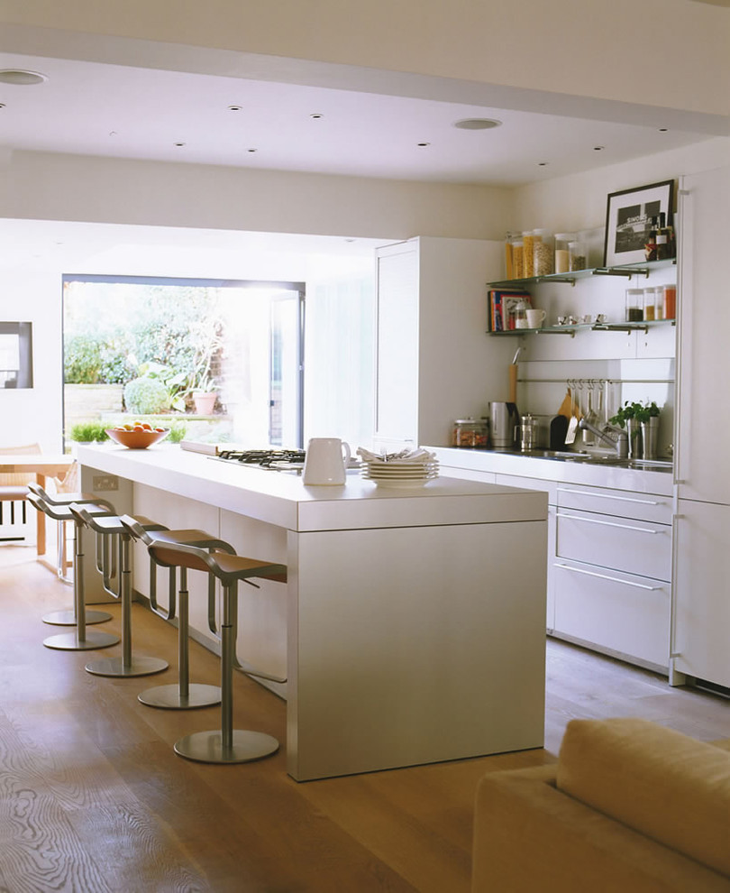 This is an example of a mid-sized contemporary galley open plan kitchen in London with flat-panel cabinets, white cabinets, white splashback, glass sheet splashback, panelled appliances, light hardwood floors and with island.