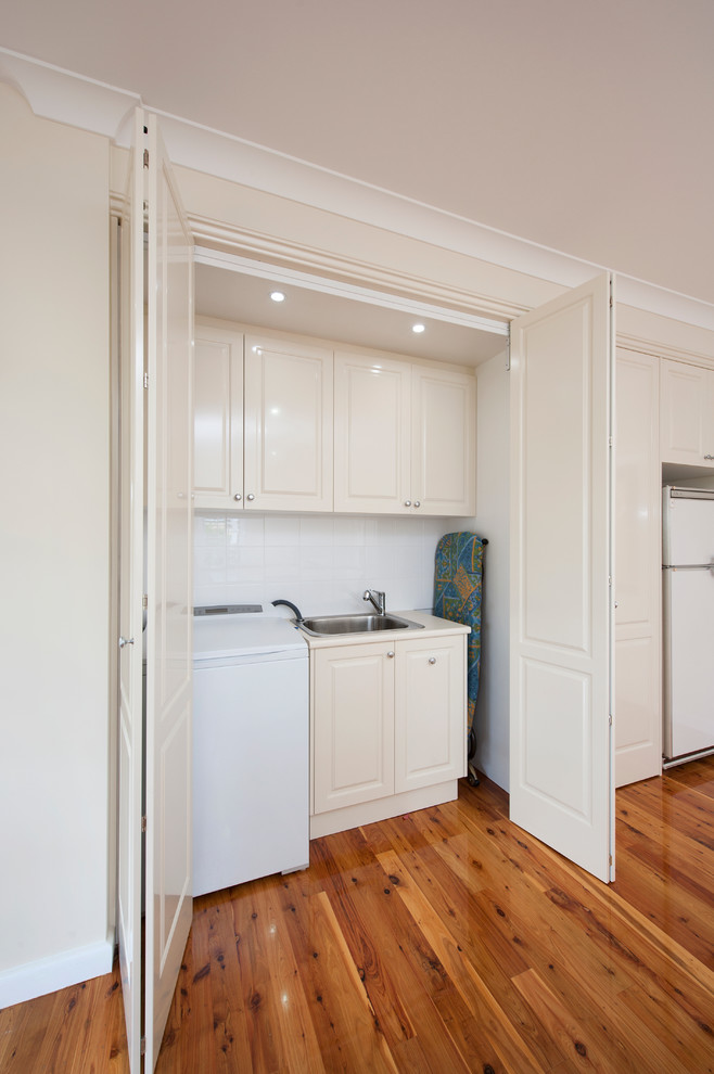 This is an example of a large traditional single-wall laundry cupboard in Melbourne with a drop-in sink, white cabinets, laminate benchtops, white walls, medium hardwood floors and an integrated washer and dryer.