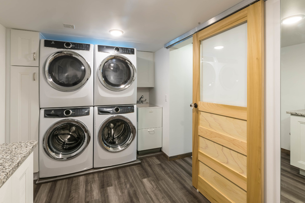 Mid-sized country l-shaped dedicated laundry room in Other with an utility sink, shaker cabinets, white cabinets, granite benchtops, white walls, vinyl floors, a stacked washer and dryer, grey floor and multi-coloured benchtop.
