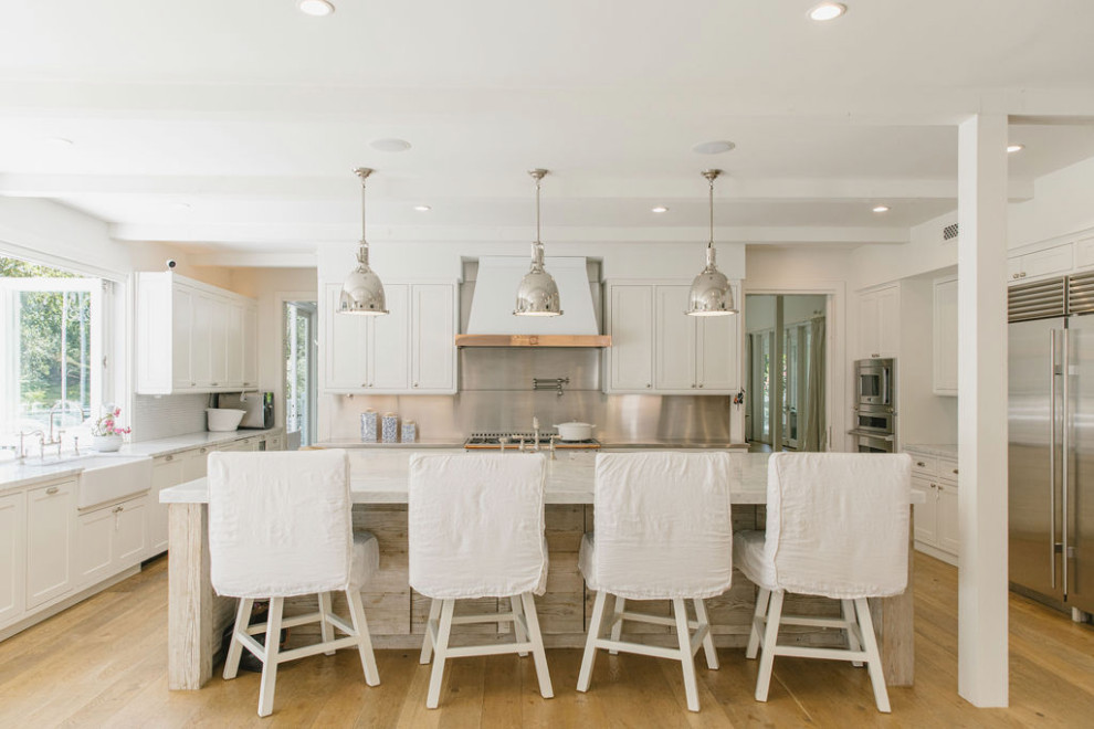 This is an example of a large country u-shaped open plan kitchen in Los Angeles with a farmhouse sink, recessed-panel cabinets, white cabinets, marble benchtops, metallic splashback, porcelain splashback, white appliances, light hardwood floors, with island, brown floor, white benchtop and wood.