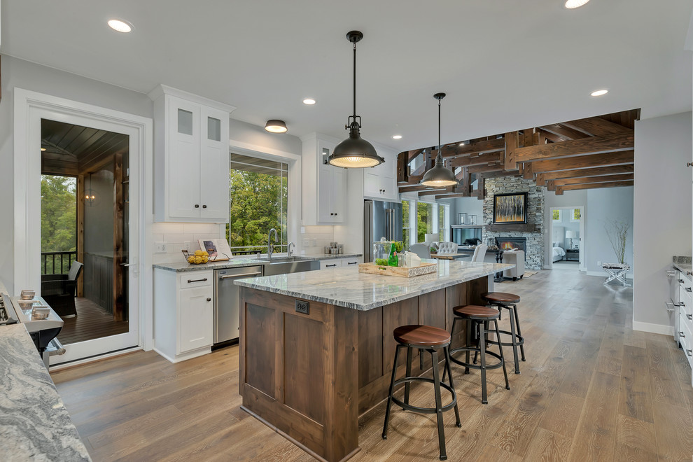 Design ideas for a mid-sized country u-shaped open plan kitchen in Minneapolis with a farmhouse sink, shaker cabinets, white cabinets, granite benchtops, white splashback, ceramic splashback, stainless steel appliances, medium hardwood floors, with island, brown floor and grey benchtop.