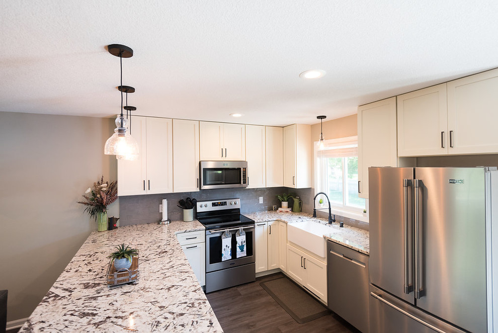 This is an example of a mid-sized country u-shaped eat-in kitchen in Minneapolis with a farmhouse sink, shaker cabinets, white cabinets, granite benchtops, grey splashback, glass tile splashback, stainless steel appliances, vinyl floors, a peninsula, brown floor and white benchtop.