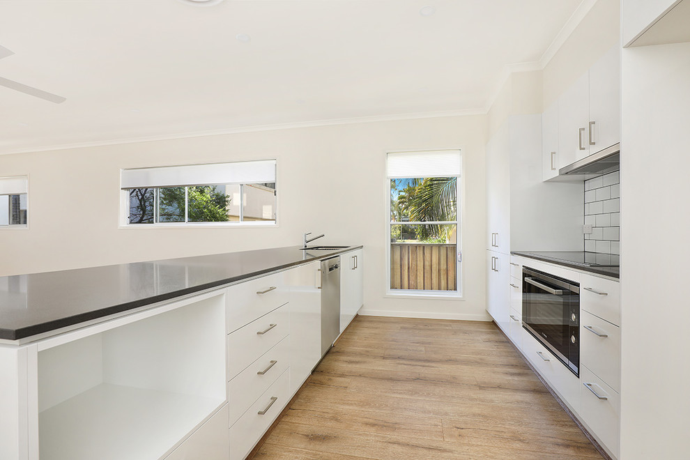 This is an example of a small beach style galley kitchen in Sunshine Coast with an undermount sink, flat-panel cabinets, white cabinets, solid surface benchtops, white splashback, subway tile splashback, stainless steel appliances, laminate floors, with island, brown floor and grey benchtop.