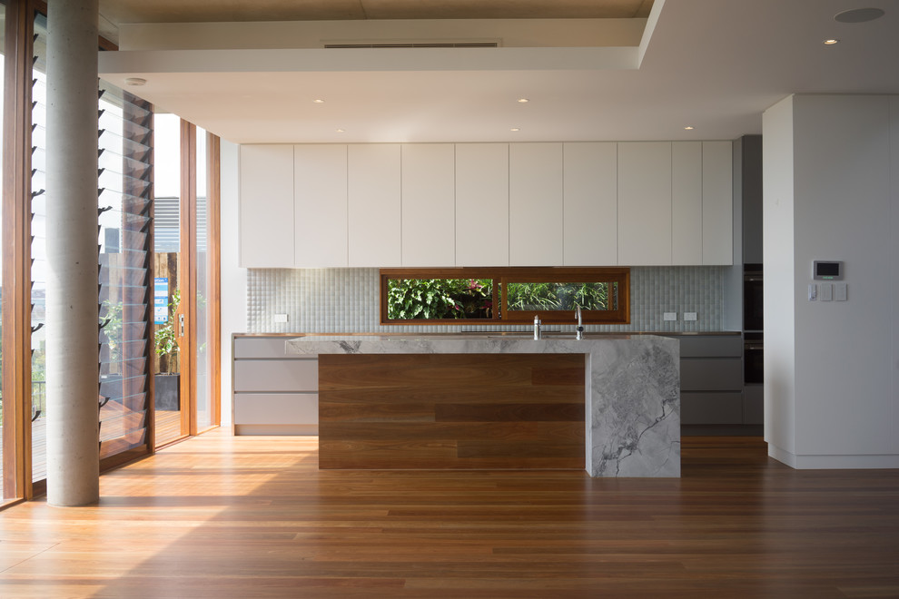 Photo of a mid-sized modern galley kitchen pantry in Brisbane with a peninsula, an undermount sink, flat-panel cabinets, white cabinets, granite benchtops, green splashback, mosaic tile splashback, black appliances, medium hardwood floors and brown floor.