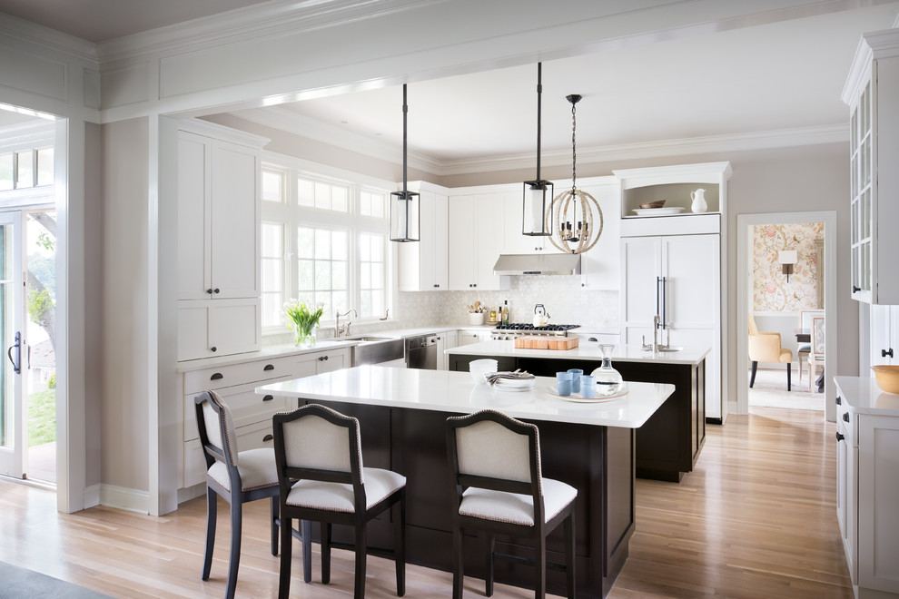 Large transitional light wood floor eat-in kitchen photo in New York with a farmhouse sink, shaker cabinets, white cabinets, solid surface countertops, white backsplash, paneled appliances and two islands