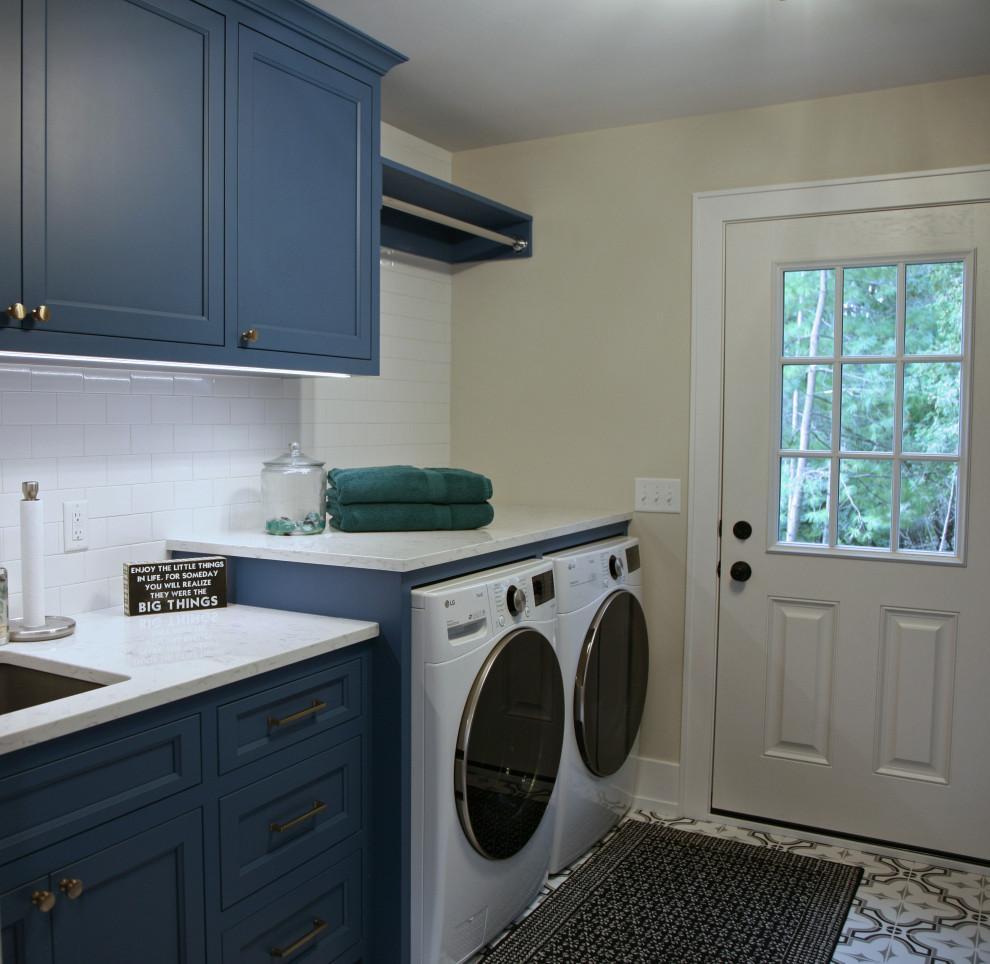 ミルウォーキーにある高級な中くらいなビーチスタイルのおしゃれな洗濯室 (ll型、アンダーカウンターシンク、シェーカースタイル扉のキャビネット、青いキャビネット、クオーツストーンカウンター、黄色いキッチンパネル、サブウェイタイルのキッチンパネル、ベージュの壁、磁器タイルの床、左右配置の洗濯機・乾燥機、グレーの床、白いキッチンカウンター) の写真