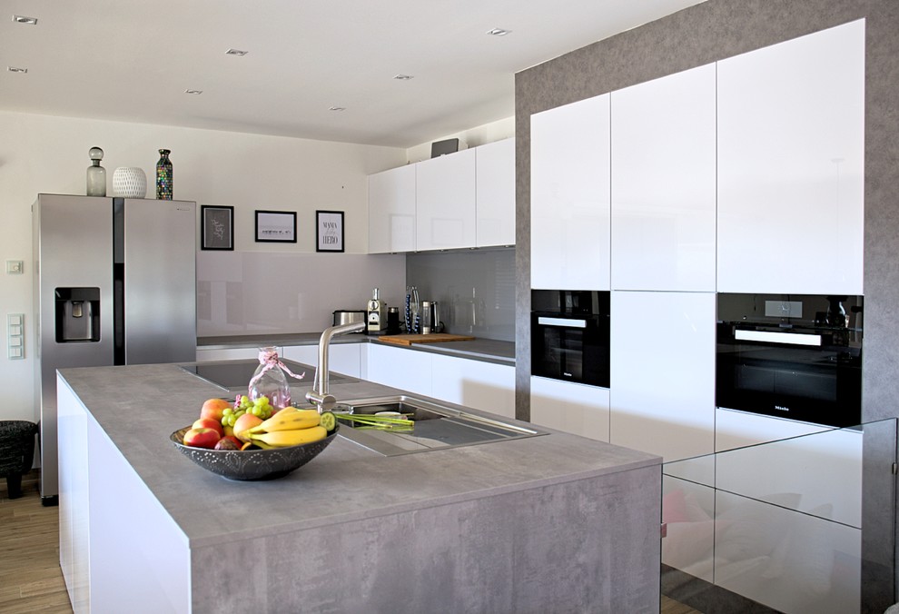 This is an example of a mid-sized industrial l-shaped separate kitchen in Berlin with white cabinets, grey splashback, glass sheet splashback, with island, a drop-in sink, flat-panel cabinets, wood benchtops, black appliances, medium hardwood floors, brown floor and grey benchtop.