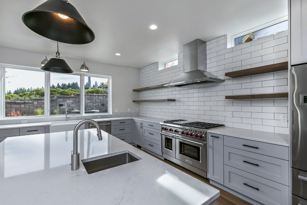 Large country l-shaped kitchen in Other with a farmhouse sink, shaker cabinets, grey cabinets, quartzite benchtops, white splashback, ceramic splashback, stainless steel appliances, light hardwood floors, with island and white benchtop.