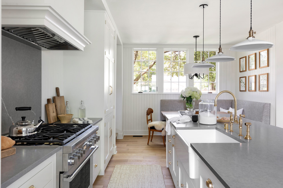 Design ideas for a mid-sized country galley open plan kitchen in Minneapolis with a farmhouse sink, shaker cabinets, white cabinets, quartz benchtops, grey splashback, engineered quartz splashback, stainless steel appliances, light hardwood floors, with island, brown floor and grey benchtop.