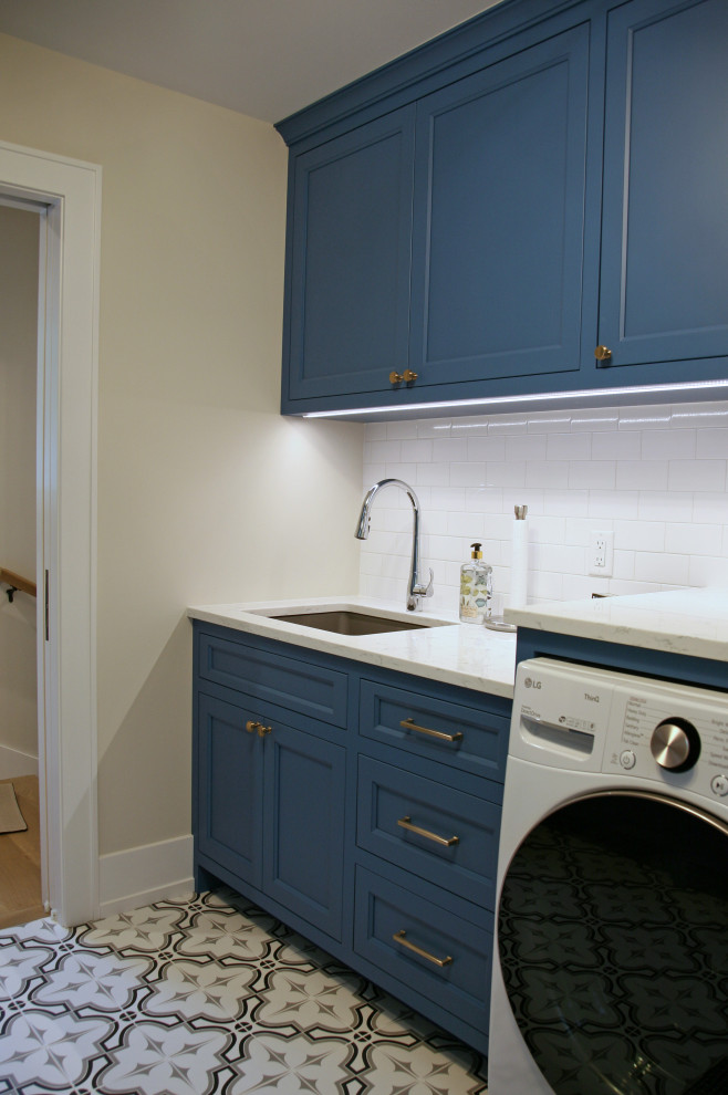 Mid-sized beach style galley dedicated laundry room in Milwaukee with an undermount sink, shaker cabinets, blue cabinets, quartz benchtops, yellow splashback, subway tile splashback, beige walls, porcelain floors, a side-by-side washer and dryer, grey floor and white benchtop.