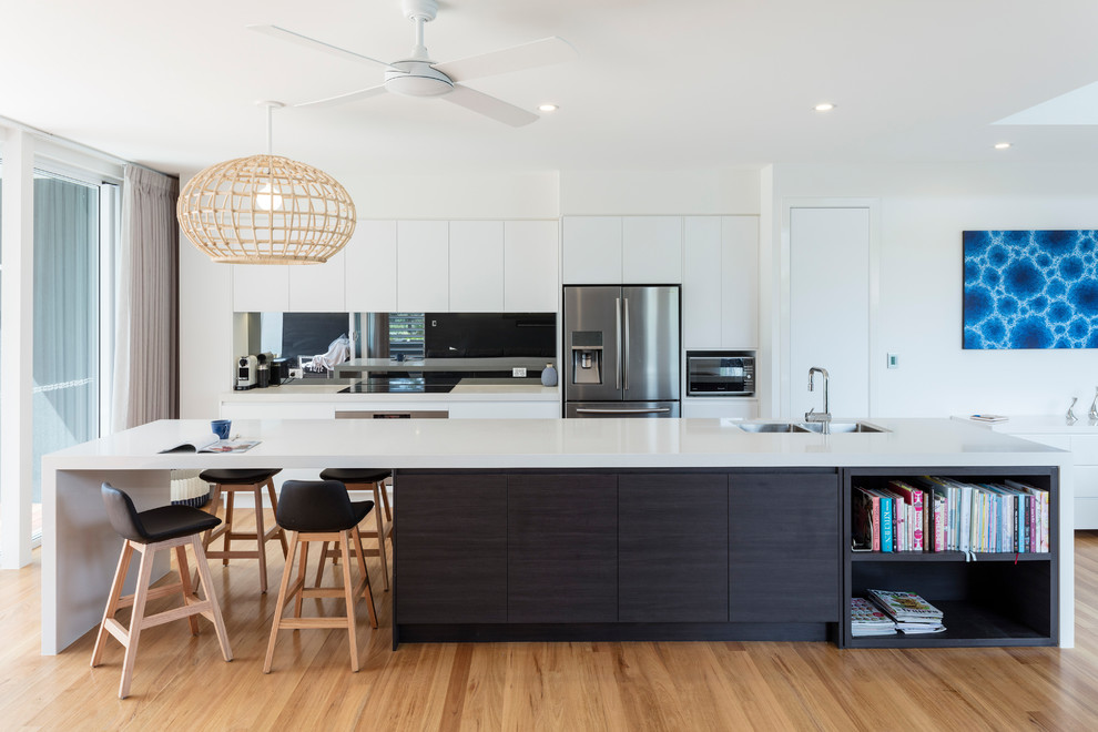 Photo of a large contemporary galley kitchen in Sunshine Coast with an undermount sink, flat-panel cabinets, white cabinets, metallic splashback, mirror splashback, stainless steel appliances, medium hardwood floors, with island, beige floor and white benchtop.