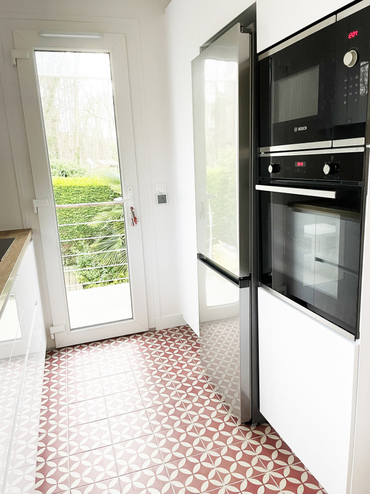パリにある高級な中くらいなミッドセンチュリースタイルのおしゃれなキッチン (アンダーカウンターシンク、フラットパネル扉のキャビネット、白いキャビネット、木材カウンター、白いキッチンパネル、ガラス板のキッチンパネル、黒い調理設備、セラミックタイルの床、アイランドなし、赤い床、茶色いキッチンカウンター) の写真