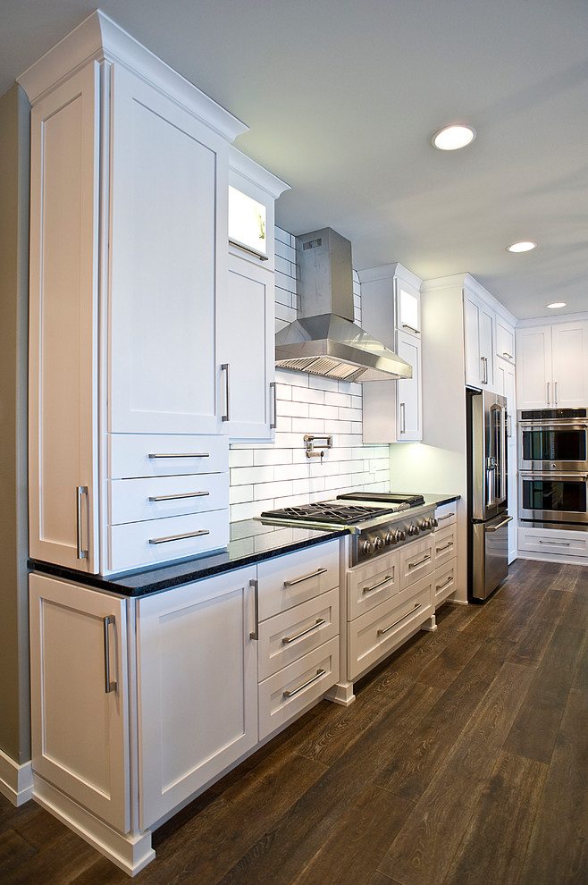 Example of a large transitional l-shaped dark wood floor and brown floor eat-in kitchen design with an undermount sink, white cabinets, granite countertops, white backsplash, subway tile backsplash, stainless steel appliances, an island and shaker cabinets