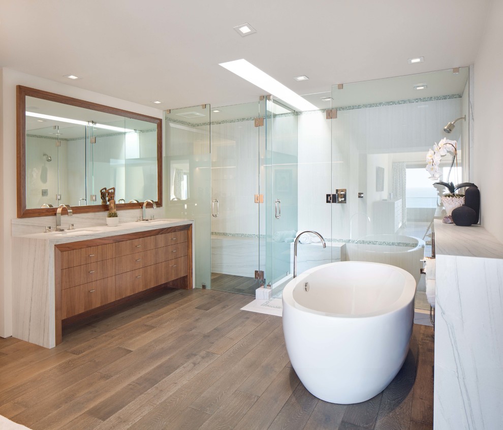 Mid-sized contemporary master bathroom in Orange County with flat-panel cabinets, a freestanding tub, white tile, marble, white walls, medium hardwood floors, an undermount sink, marble benchtops, medium wood cabinets, a corner shower, brown floor, a hinged shower door and white benchtops.