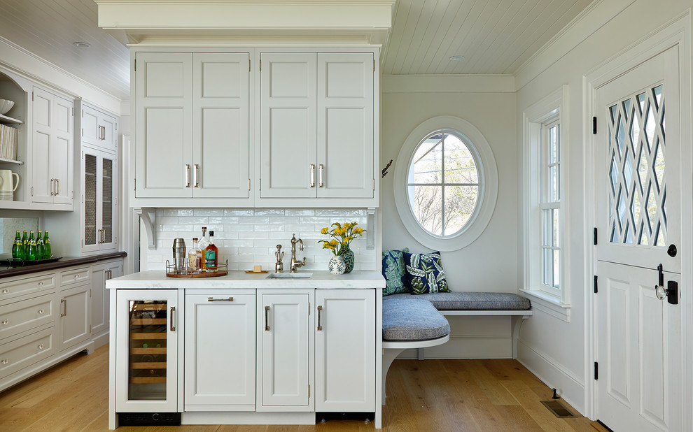 This is an example of a large transitional single-wall eat-in kitchen in New York with a farmhouse sink, shaker cabinets, white cabinets, quartz benchtops, white splashback, ceramic splashback, stainless steel appliances, light hardwood floors, with island, brown floor and white benchtop.