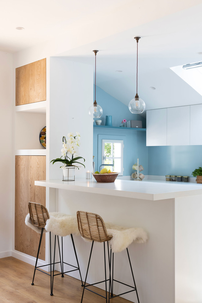 Photo of a beach style kitchen in Bordeaux with flat-panel cabinets, white cabinets, blue splashback, a peninsula, white benchtop and light hardwood floors.