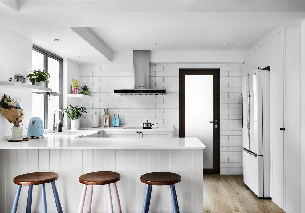 This is an example of a contemporary u-shaped kitchen in Singapore with a farmhouse sink, shaker cabinets, white cabinets, white splashback, subway tile splashback, white appliances, light hardwood floors, a peninsula and white benchtop.