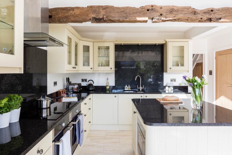 Large transitional l-shaped open plan kitchen in West Midlands with a double-bowl sink, glass-front cabinets, white cabinets, granite benchtops, black splashback, laminate floors, with island, black benchtop and exposed beam.