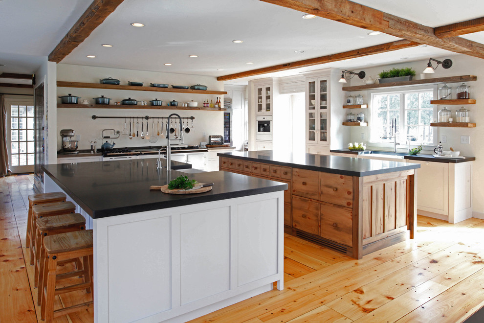 Photo of a large country u-shaped kitchen in New York with a farmhouse sink, shaker cabinets, granite benchtops, medium hardwood floors, multiple islands, white cabinets, white appliances and brown floor.