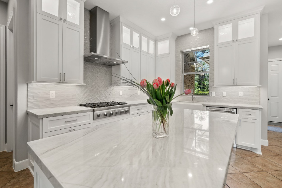 This is an example of a mid-sized transitional l-shaped eat-in kitchen in Houston with a farmhouse sink, recessed-panel cabinets, white cabinets, quartzite benchtops, grey splashback, ceramic splashback, stainless steel appliances, porcelain floors, with island, beige floor and grey benchtop.
