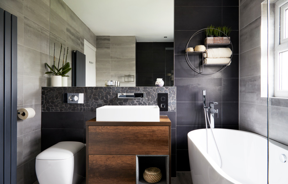 Mid-sized contemporary 3/4 bathroom in Surrey with flat-panel cabinets, medium wood cabinets, a freestanding tub, a shower/bathtub combo, a wall-mount toilet, gray tile, porcelain tile, a vessel sink and brown benchtops.