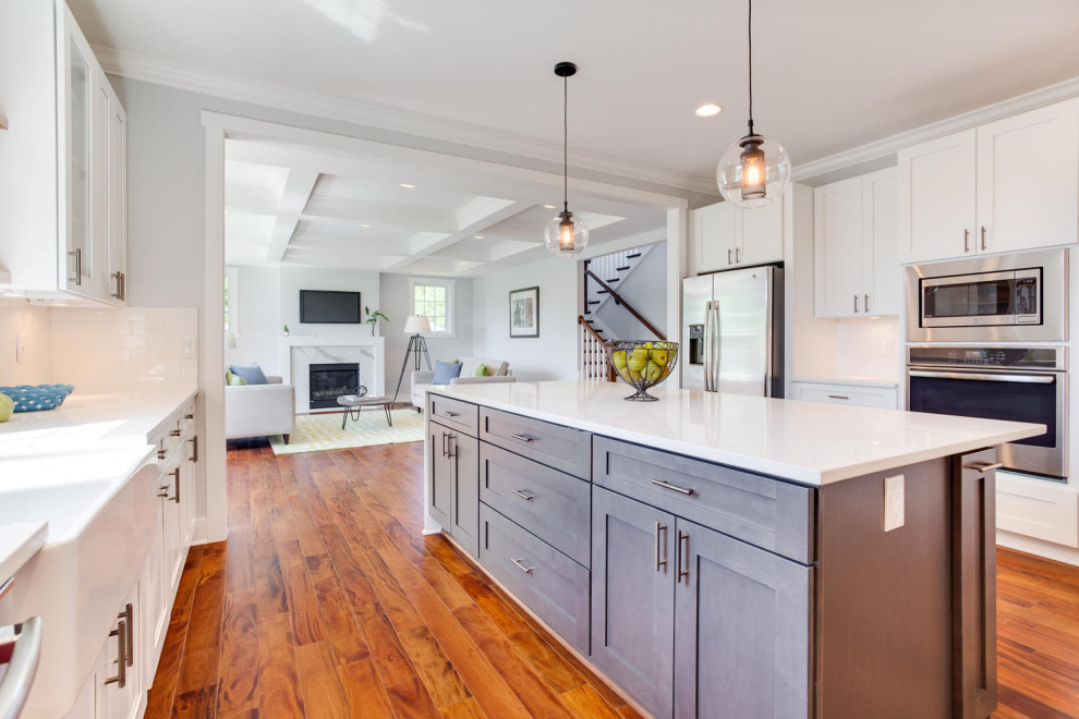 Design ideas for a traditional u-shaped open plan kitchen in DC Metro with a belfast sink, shaker cabinets, white cabinets, engineered stone countertops, white splashback, ceramic splashback, stainless steel appliances, medium hardwood flooring, an island and white worktops.