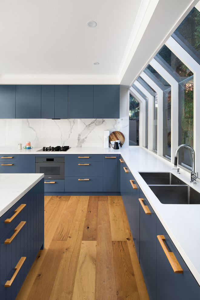 Design ideas for a large contemporary u-shaped open plan kitchen in Melbourne with blue cabinets, marble benchtops, white splashback, marble splashback, with island, a drop-in sink, flat-panel cabinets, stainless steel appliances, medium hardwood floors, brown floor and white benchtop.