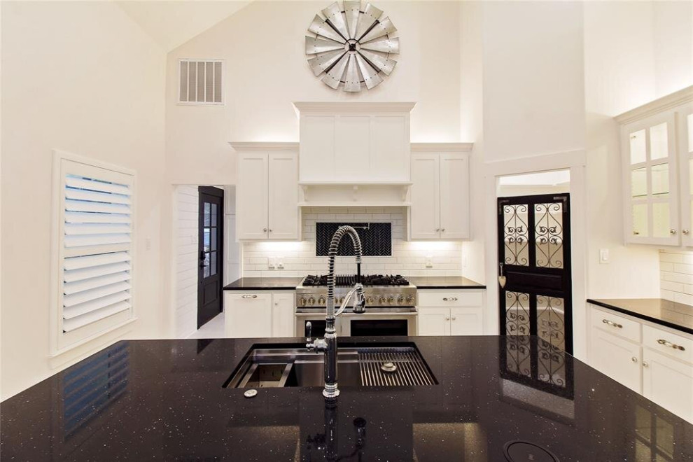 This is an example of a mid-sized transitional l-shaped eat-in kitchen in Houston with a triple-bowl sink, shaker cabinets, white cabinets, quartzite benchtops, white splashback, ceramic splashback, stainless steel appliances, porcelain floors, with island, multi-coloured floor, black benchtop and coffered.