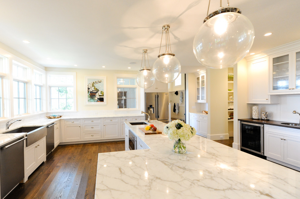 This is an example of a beach style l-shaped open plan kitchen in Boston with a farmhouse sink, recessed-panel cabinets, white cabinets, marble benchtops, white splashback, subway tile splashback, stainless steel appliances, medium hardwood floors, with island and brown floor.