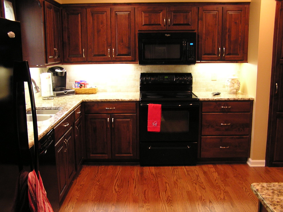 This is an example of a mid-sized traditional l-shaped eat-in kitchen in Kansas City with raised-panel cabinets, granite benchtops, black appliances, with island, a double-bowl sink, medium wood cabinets, beige splashback, stone tile splashback, medium hardwood floors and brown floor.