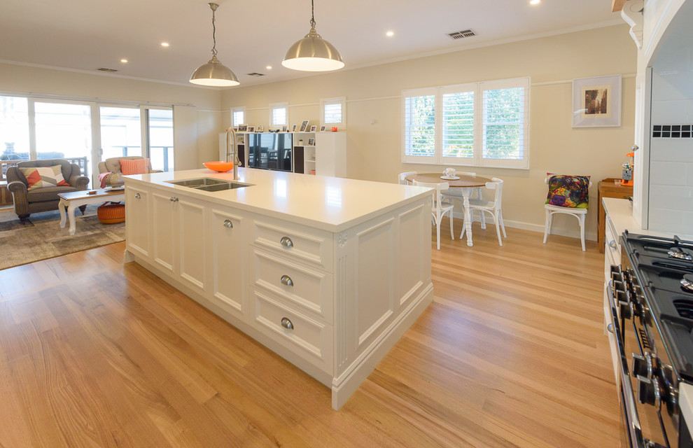 Photo of a large transitional l-shaped open plan kitchen in Melbourne with an undermount sink, recessed-panel cabinets, white cabinets, quartz benchtops, white splashback, subway tile splashback, black appliances, light hardwood floors and with island.