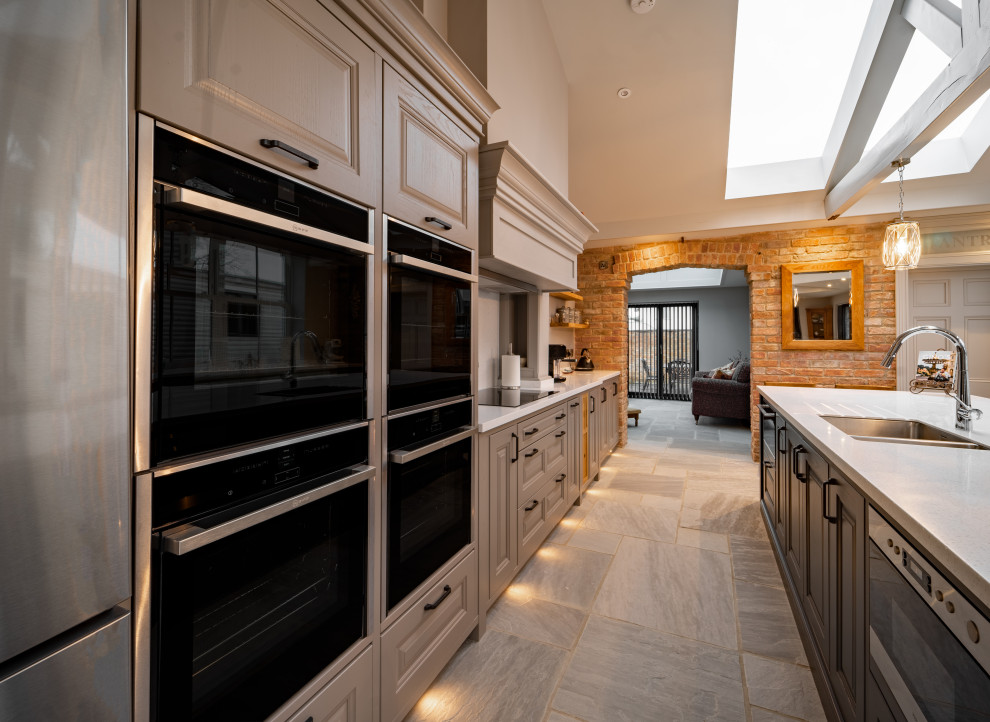 This is an example of a large traditional galley open plan kitchen in Other with an integrated sink, raised-panel cabinets, grey cabinets, composite countertops, white splashback, black appliances, ceramic flooring, an island, grey floors, white worktops and a vaulted ceiling.