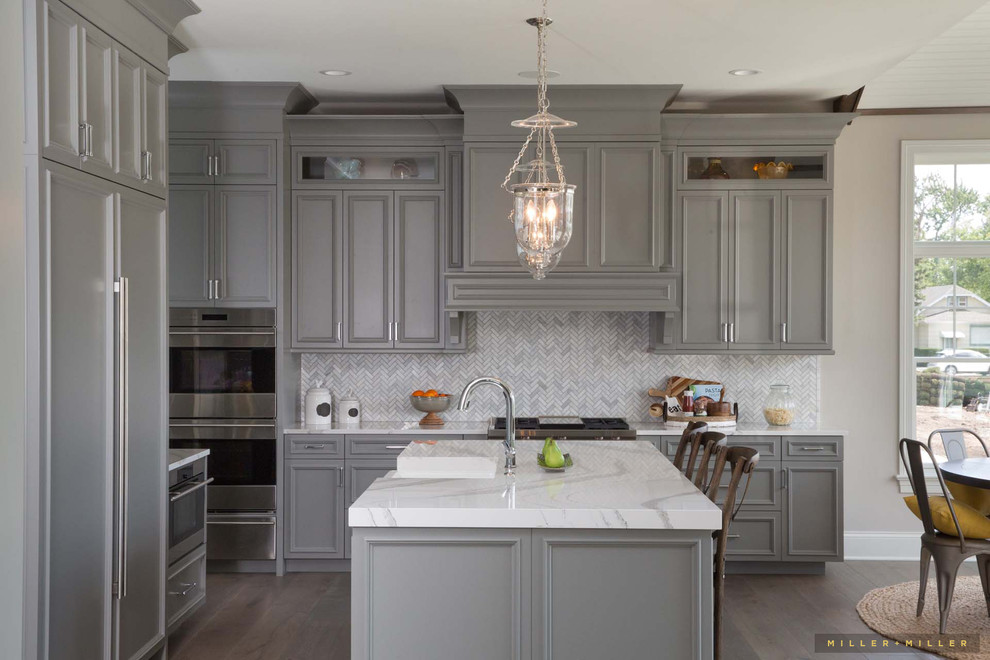 Photo of a large country l-shaped kitchen in Chicago with a farmhouse sink, beaded inset cabinets, grey cabinets, marble benchtops, grey splashback, marble splashback, panelled appliances, medium hardwood floors, with island, brown floor and white benchtop.
