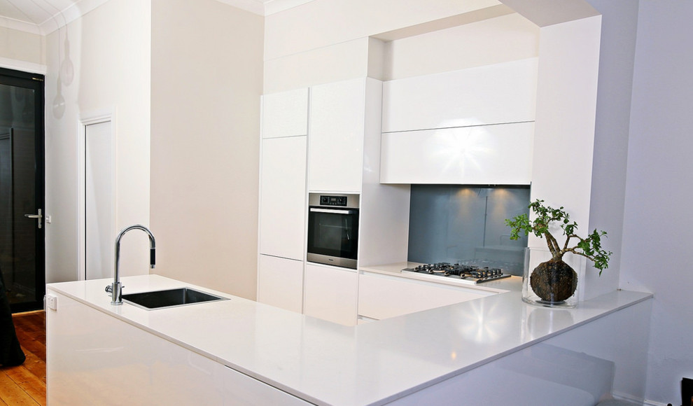 Photo of a contemporary u-shaped eat-in kitchen in Sydney with a single-bowl sink, flat-panel cabinets, white cabinets, granite benchtops, blue splashback, glass sheet splashback, stainless steel appliances, medium hardwood floors and no island.
