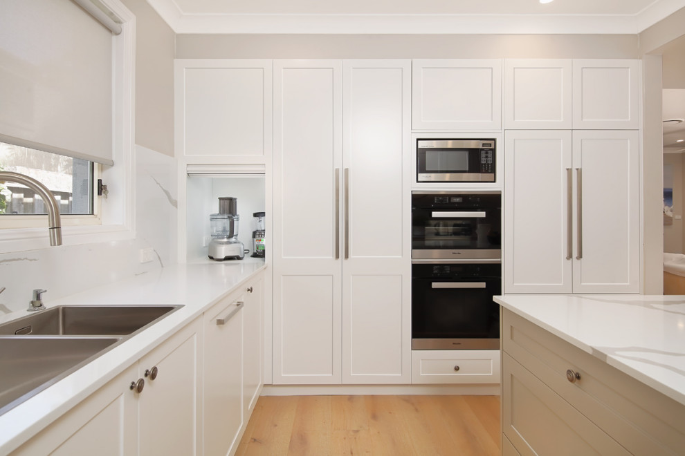 This is an example of a large modern u-shaped eat-in kitchen in Sydney with an integrated sink, raised-panel cabinets, white cabinets, quartz benchtops, white splashback, stone slab splashback, panelled appliances, dark hardwood floors, with island, brown floor and white benchtop.