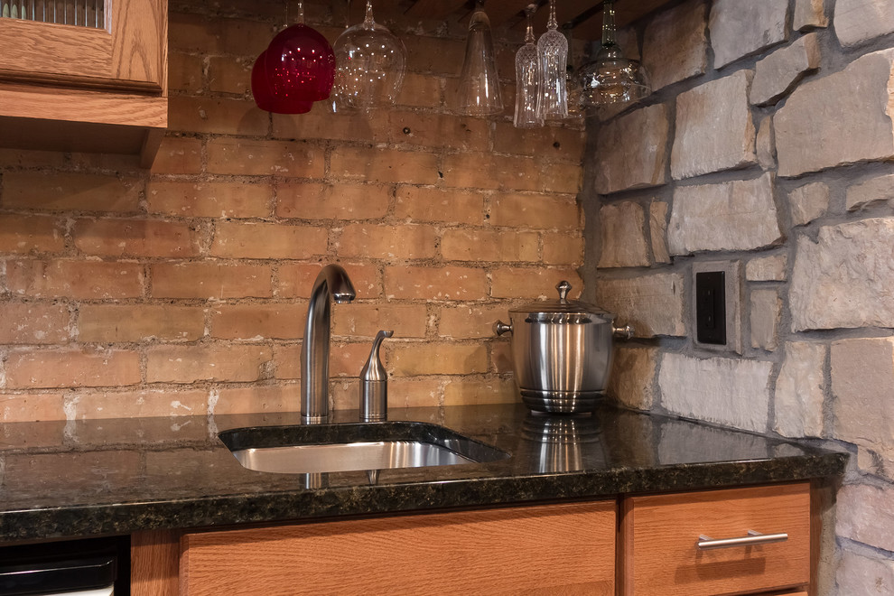 This is an example of a mid-sized transitional single-wall wet bar in Minneapolis with an undermount sink, recessed-panel cabinets, medium wood cabinets, granite benchtops, beige splashback, brick splashback, dark hardwood floors, black floor and black benchtop.