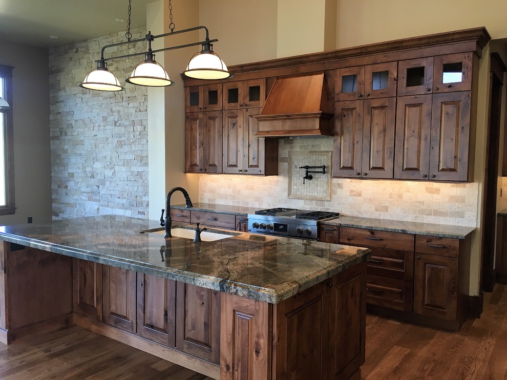 Photo of a large country l-shaped kitchen in Denver with an undermount sink, raised-panel cabinets, brown cabinets, granite benchtops, beige splashback, terra-cotta splashback, stainless steel appliances, medium hardwood floors and with island.
